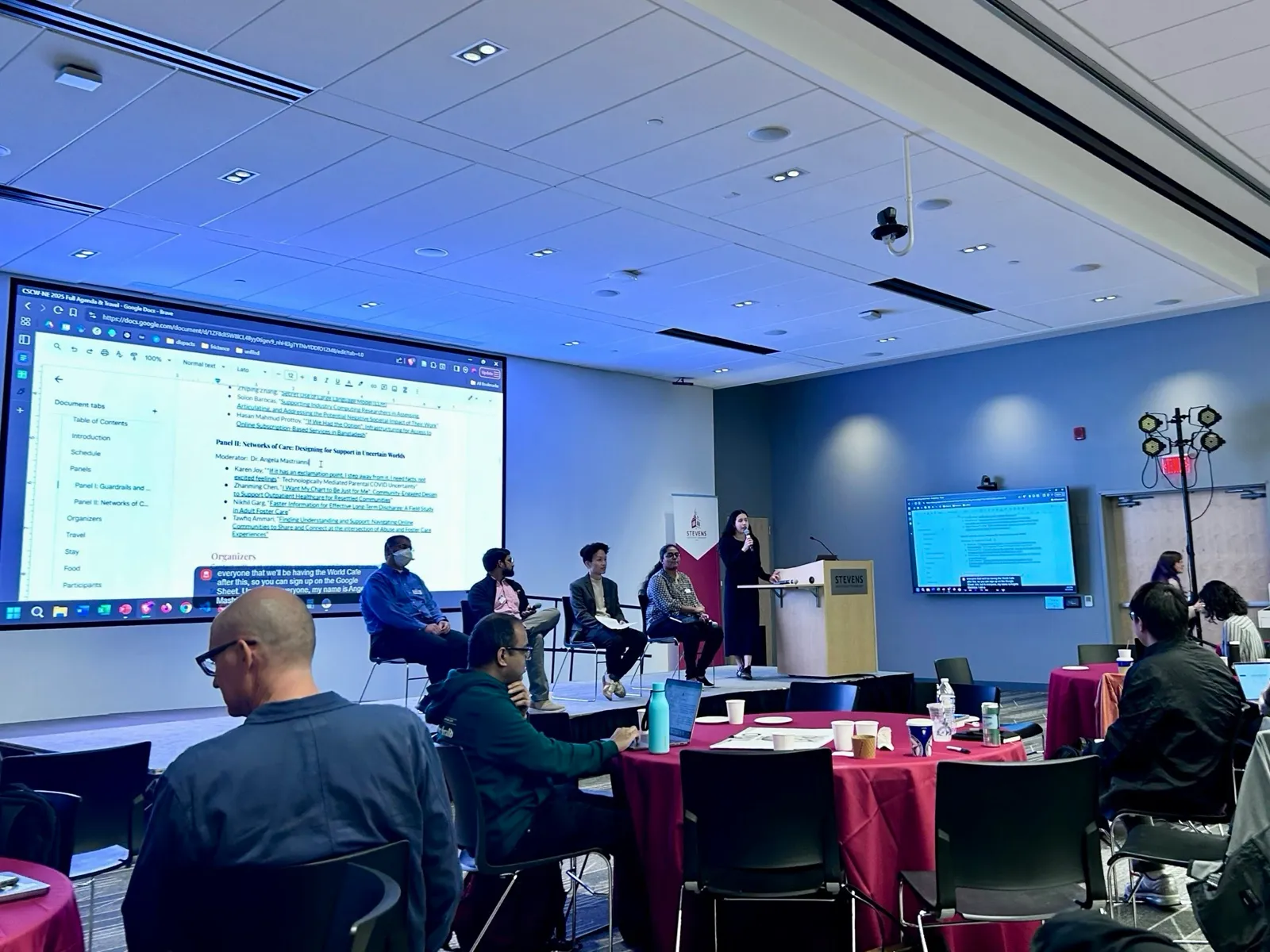 Four panelists seated on a stage, Zhanming Chen third from left, with a moderator at a lectern, a projected agenda and attendees at round tables.